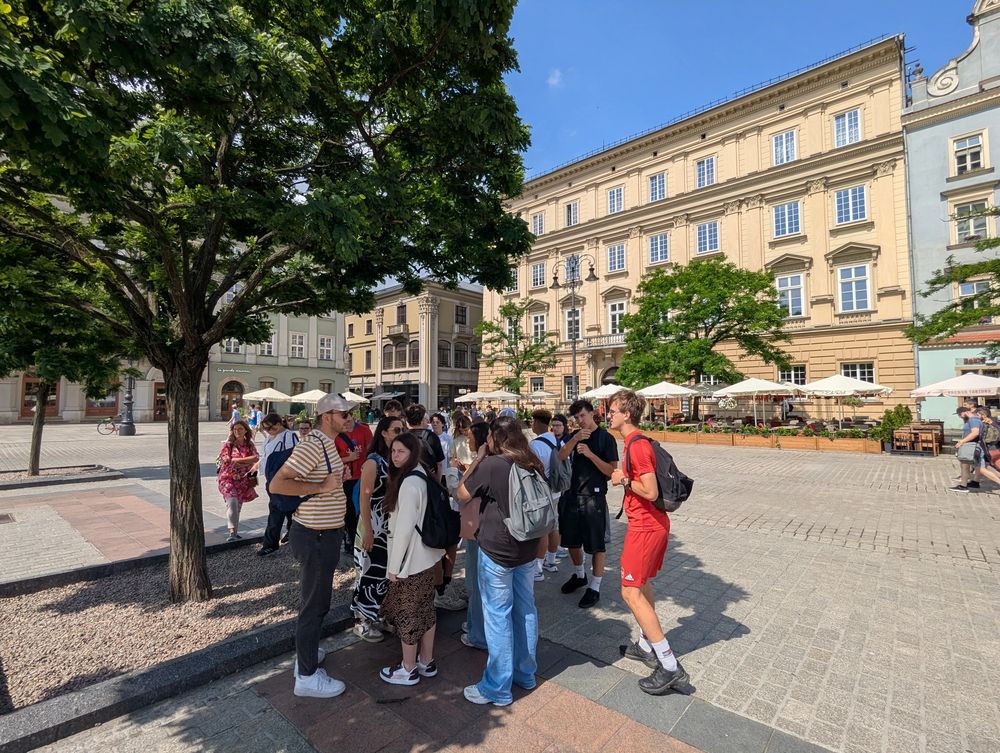 Le groupe discute sous un grand arbre, façades anciennes et terrasses derrière.