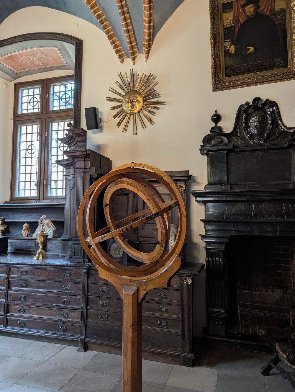 Grande sphère armillaire en bois sur pied, soleil doré au mur et portrait ancien.