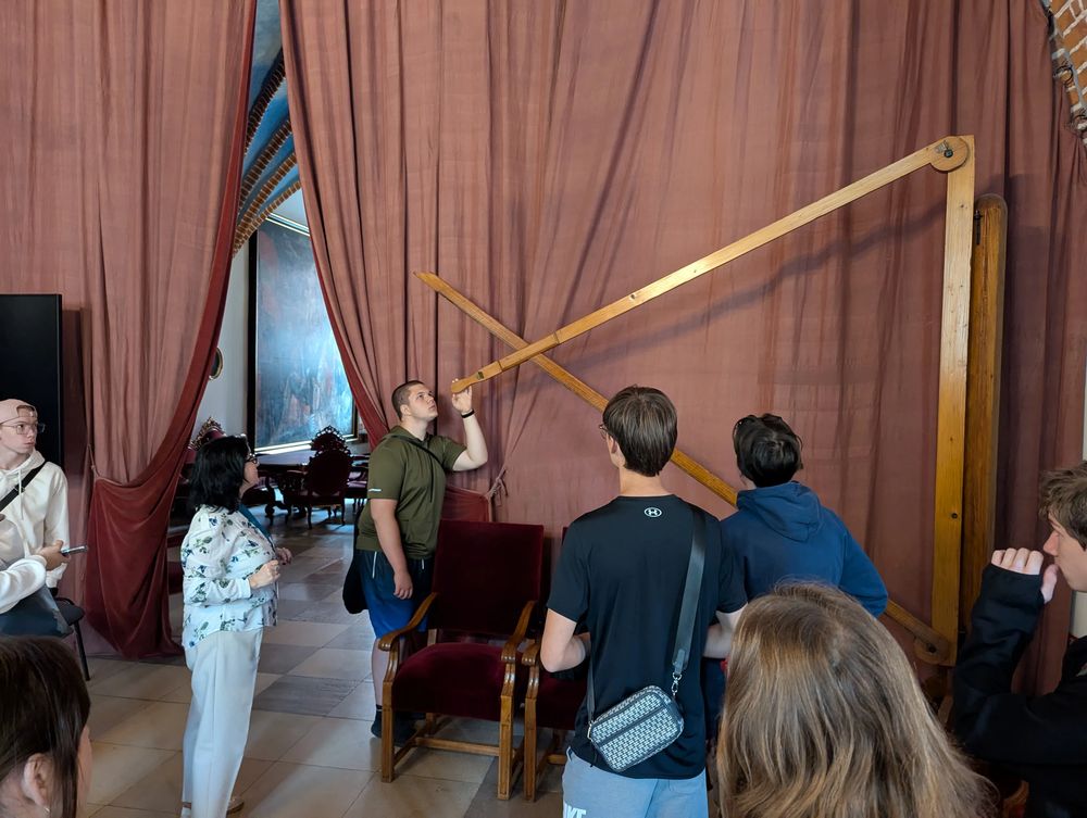 Des visiteurs observent un grand instrument de mesure en bois sur un mur tendu de tissu rouge.