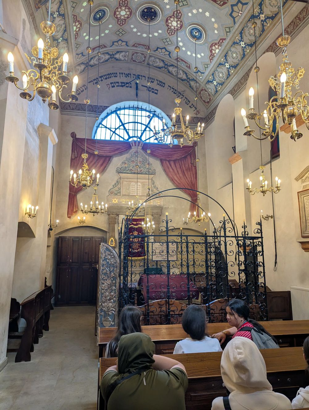 Intérieur d'une synagogue : plafond peint, estrade (bima) entourée d'une grille en fer forgé, inscriptions en hébreu ; le groupe est assis.