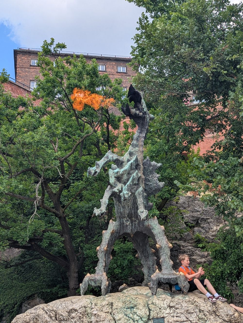 Statue en bronze du dragon de Wawel crachant une flamme, au pied de la colline, entourée d'arbres.