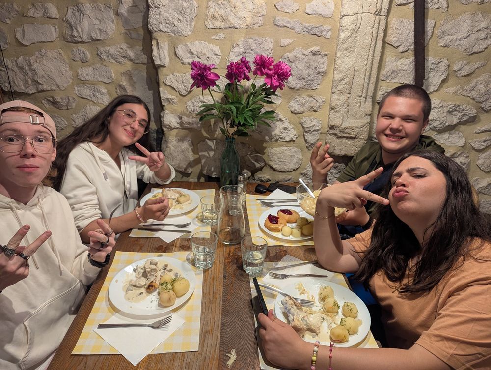 Quatre convives attablés font le geste de la victoire, devant des assiettes de poulet en sauce et pommes de terre.
