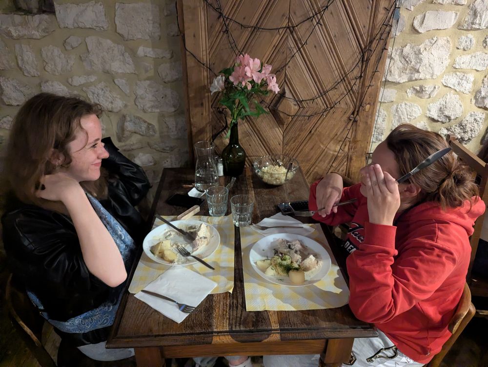 Deux convives se font face à une petite table, assiettes de poulet aux pommes de terre et fleurs au centre.