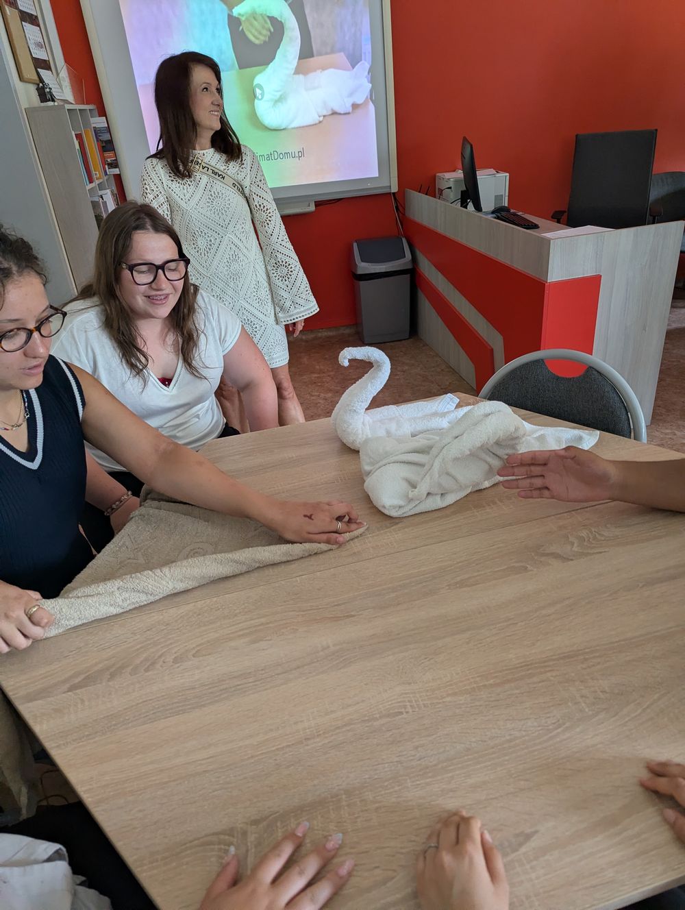 Une enseignante près d'un écran montrant un cygne en serviette, avec un cygne plié posé sur la table.