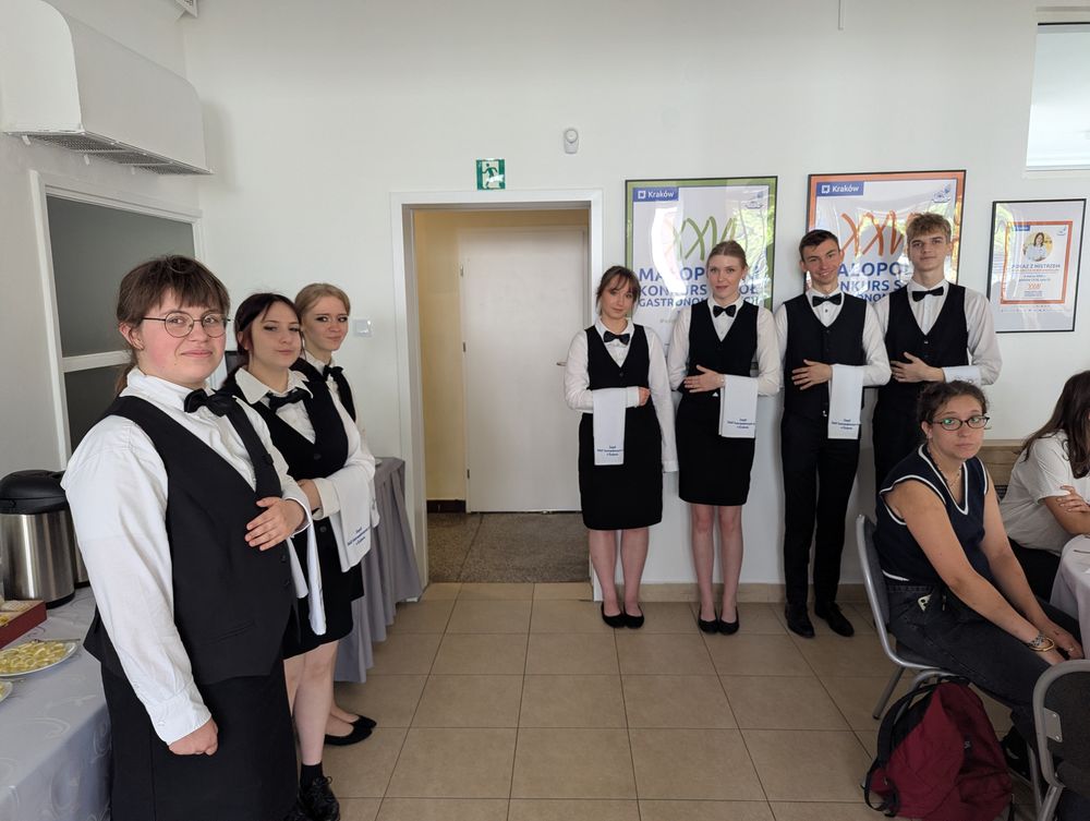 Des élèves de l'école en tenue de service noir et blanc, la serviette sur le bras, alignés.