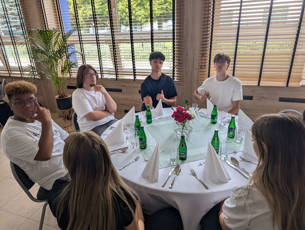 Des convives autour d'une table ronde dressée : nappe blanche, serviettes pliées et fleurs.