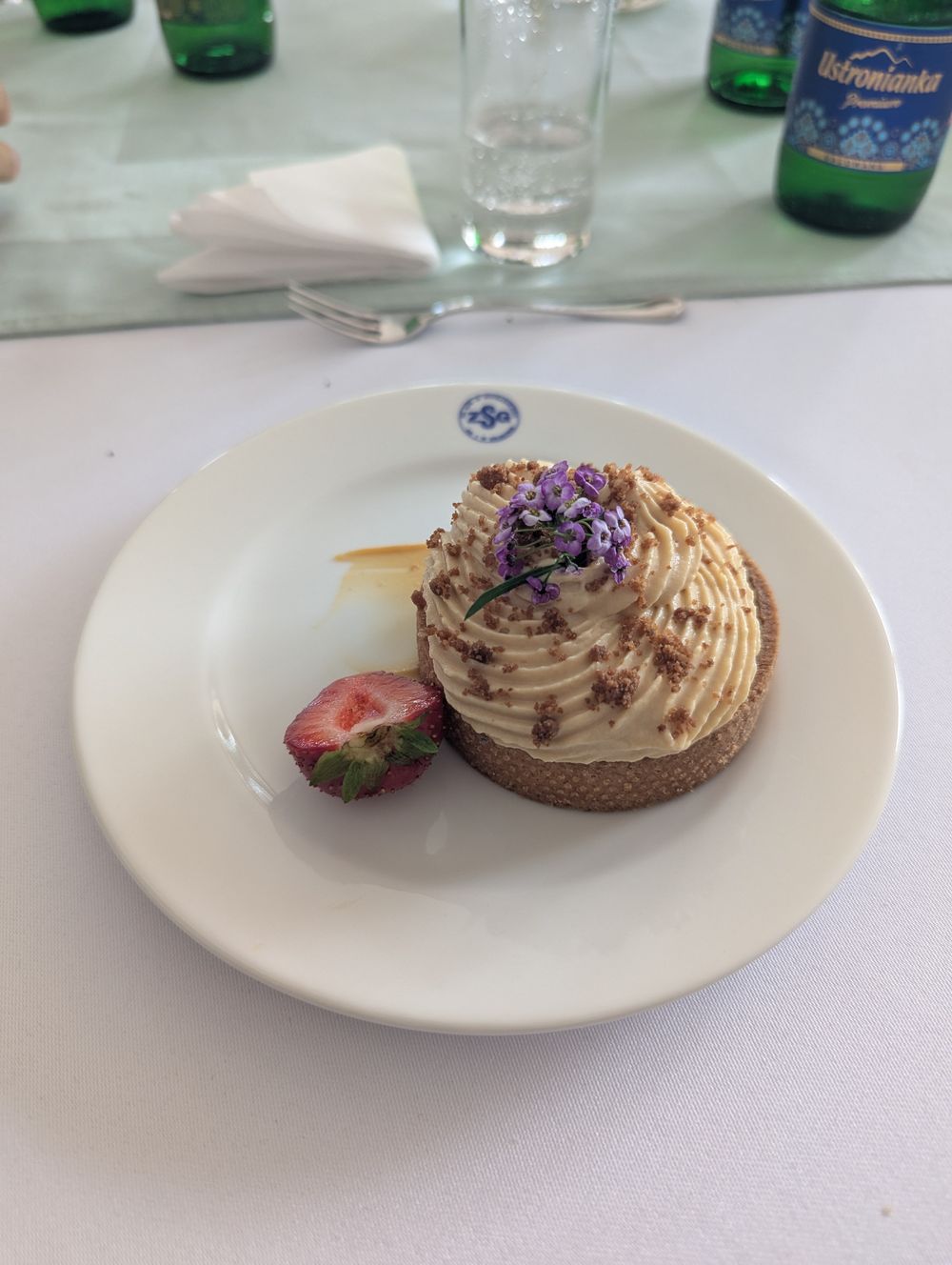 Tarte aux pommes surmontée d'une mousse caramel et d'éclats de spéculoos, décorée d'une demi-fraise et de fleurs comestibles, sur une assiette au logo ZSG.