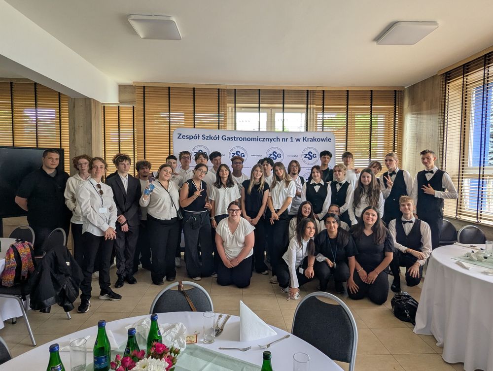 Grande photo de groupe de la délégation et des élèves de l'école devant la bannière « Zespół Szkół Gastronomicznych nr 1 w Krakowie », dans la salle dressée pour le déjeuner.