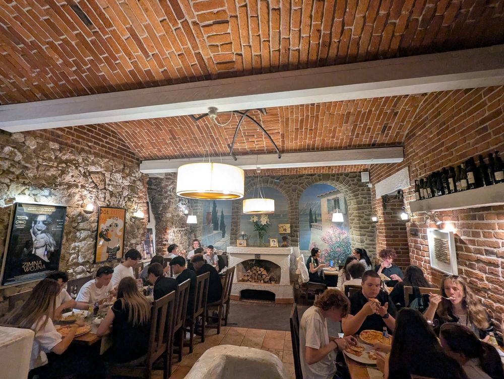 Vue de la salle voûtée en brique du restaurant italien Soprano : les convives dînent à de longues tables, parmi les fresques méditerranéennes et une cheminée en pierre.