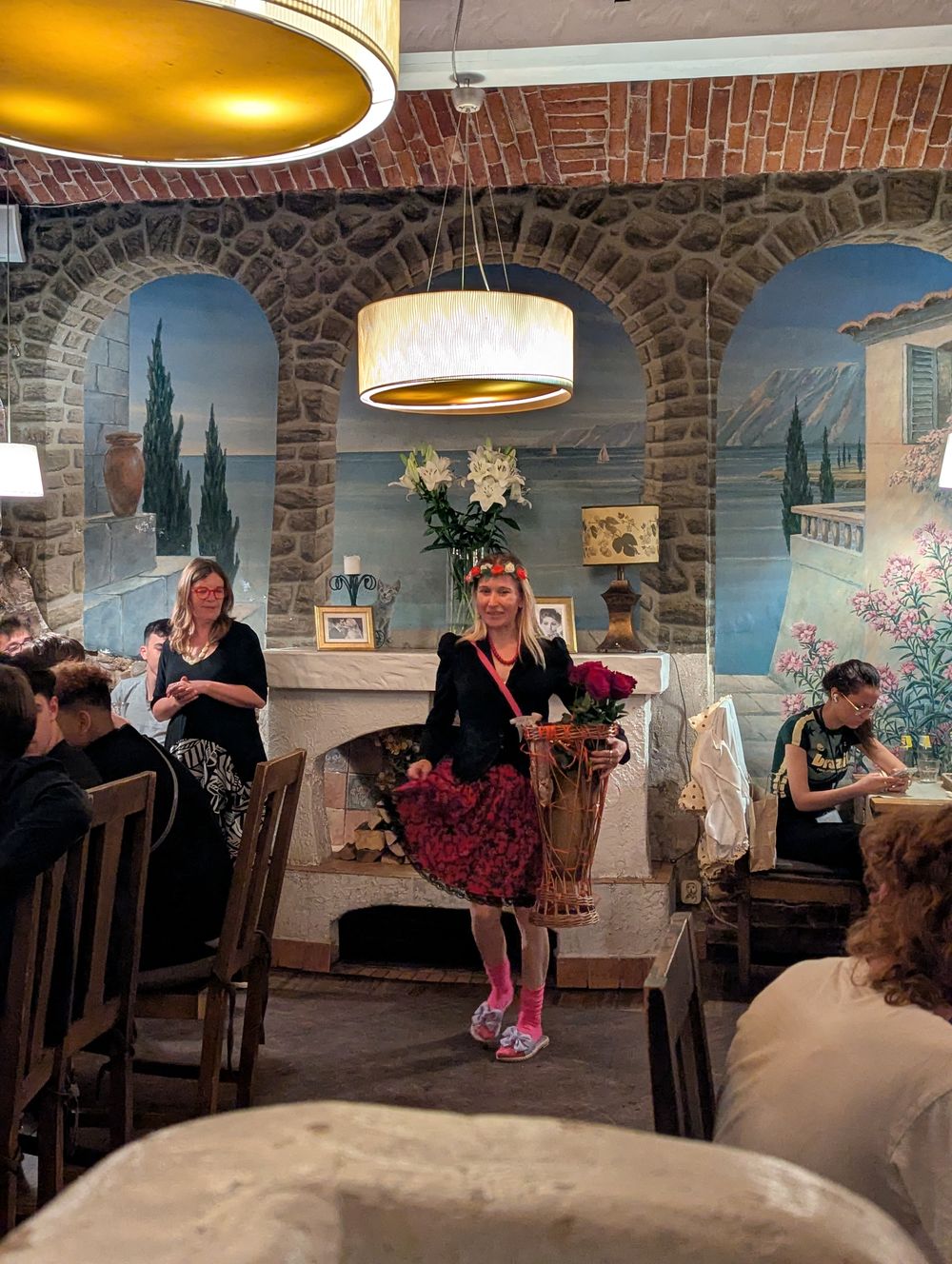 Une femme en habit folklorique polonais, couronne de fleurs et panier de roses rouges, passe entre les tables d'un restaurant aux fresques méditerranéennes.