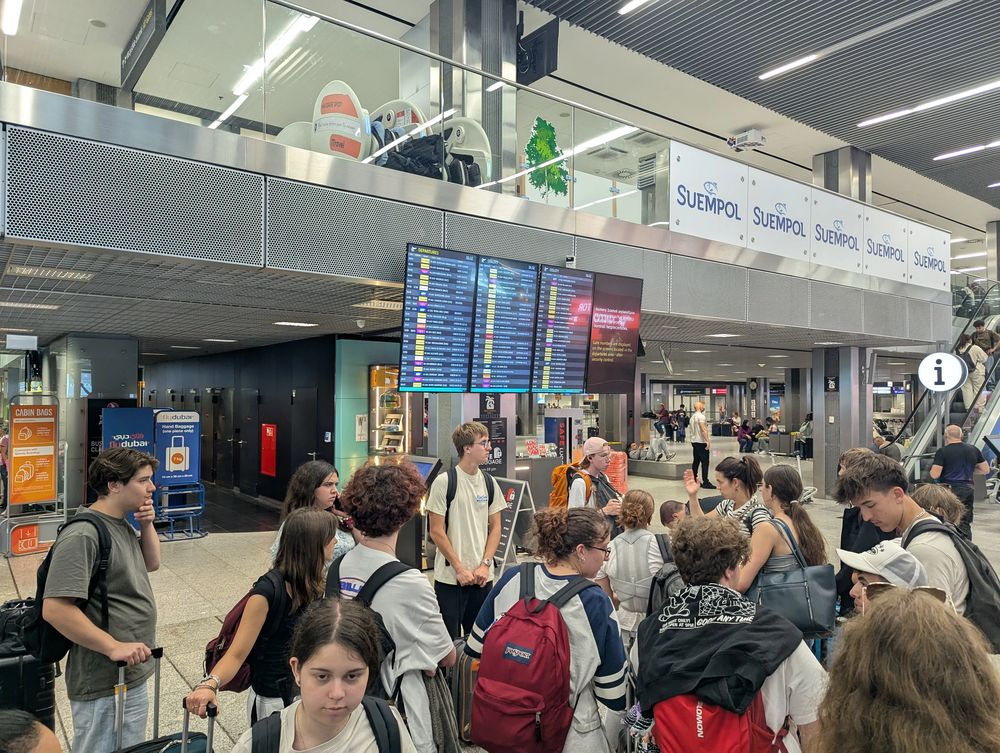 Dans l'aérogare, le groupe patiente devant le tableau des départs avant l'embarquement.